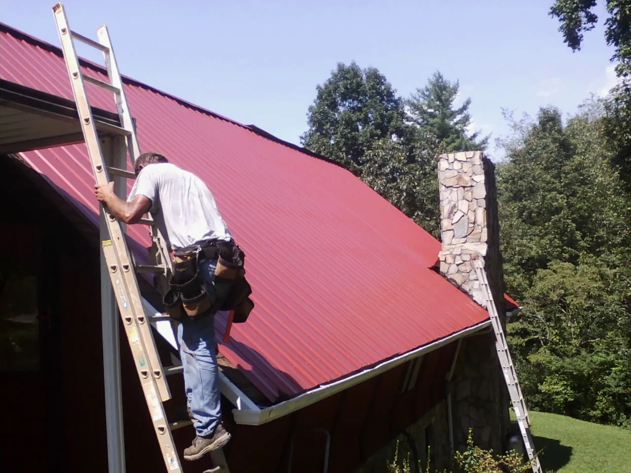 Roofer on a ladder beside a red metal roof and stone chimney — Soffit & Fascia Repair by Roofing Brothers in Avalon